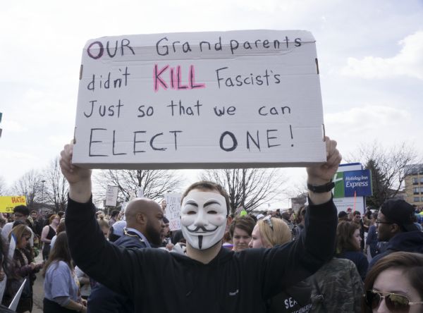 A man wearing a Guy Fawkes mask is holding up a sign which reads: "Our Grandparents didn't Kill fascist's [sic] just so that we can elect one!" He is standing in front of a crowd of protesters outside the Donald Trump rally.