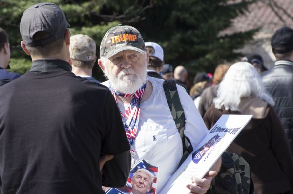 An elderly man is standing in a crowd of people waiting to enter the Donald Trump rally. He is wearing a camouflage hat with Trump's campaign slogan on it: "Make America Great Again." A megaphone with Trump's image is hanging around his neck on an American flag lanyard. He is holding a posterboard with another cut-out Trump megaphone and instructions on how to assemble it. The poster reads: "Let Them Hear You."