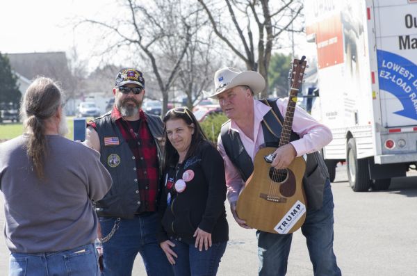Two men and a woman are posing for a photograph outside the Donald Trump rally. The man on the left is wearing a hat and vest marking him as a disabled veteran of the 82nd Airborne Division. The man on the right is wearing a cowboy hat with a Trump pin on it and is holding his guitar with a Trump bumper sticker attached to it. There is a woman standing between the men who is wearing a hoodie with numerous pro-Trump or anti-Hillary buttons pinned on it. A man in the foreground is taking the picture with a smart phone.