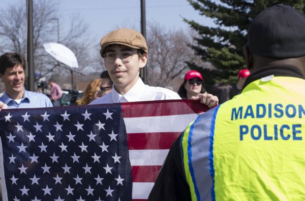 A young man in a flat cap and glasses is holding up an American flag. He is standing in a crowd of Trump supporters waiting to enter the rally. An African American man wearing a Madison Police uniform is in the foreground.