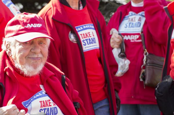 An elderly man is sitting and posing for the camera outside the Donald Trump rally. He is wearing a red "AARP" cap, a red jacket, and a red shirt which reads: "Social Security, Take a Stand." Other people standing around him are wearing the same jacket and shirt.