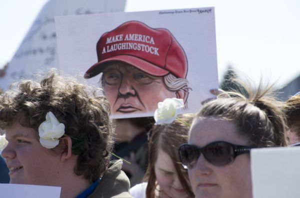 A person standing in a crowd of people protesting the rally is holding up a sign with an image of Donald Trump wearing a red cap reading: "Make America a Laughingstock." Two people in front are wearing white roses in their hair.