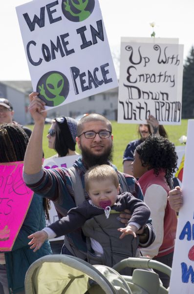 A man is holding his political poster in his left hand, which bears images of cartoon aliens and reads: "We Come in Peace." His right hand is steadying a baby in a baby carrier on his chest. The man is surrounded by men and women holding up their own political signs protesting Donald Trump. One sign in the back reads: "Up with Empathy, Down with Apathy." It has a white rose attached to it.