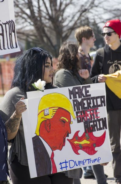 A young woman is holding a sign which reads: "Make America Hate Again," along with the hashtag: "#DumpTrump." It includes an image of Donald Trump breathing fire with dollar signs, wearing a wig with a tag stating "Made in China," and wearing a Hitler moustache. A white rose is attached to the sign. The woman is surrounded by other young men and women protesting outside the Donald Trump rally.