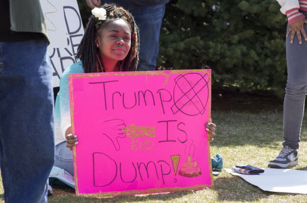 A young African American woman is sitting on the grass and holding a pink sign which reads: "Trump is Dump!" She is wearing a white rose in her hair. Other people are standing around her.