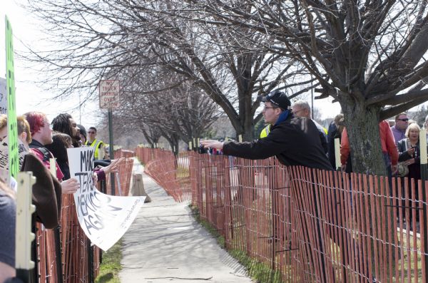 Temporary fences are along both sides of a narrow sidewalk outside the Donald Trump rally. Behind the fence on the left side are several protesters holding signs, one reading: "Make Texas Mexico Again." Trump supporters are standing behind the other fence on the right waiting to enter the rally. One of them, a young man, is leaning over the fence to address the protesters. Police are standing on both sides of the fence watching both groups.