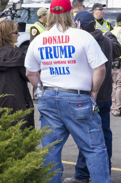View from the back of a man wearing blue jeans, a red hat, and a white t-shirt which reads: "Donald Trump Finally Someone with Balls." Two other people are standing near him, and four policemen are standing in the background next to parked cars.