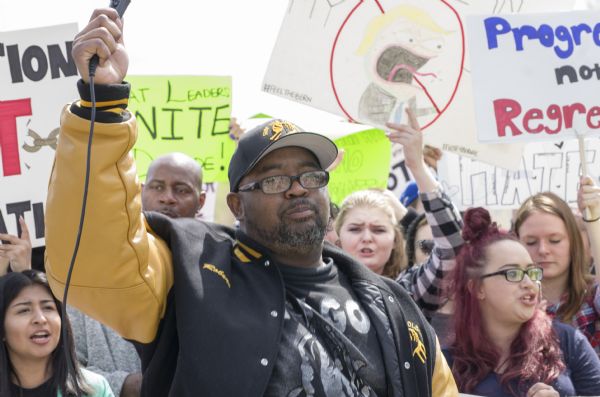 Robert Grimes is holding up a microphone and leading a group of protesters in a chant outside the Donald Trump Rally. He is wearing a jacket and hat, both of which read "Stop the Violence," and include the logo of that movement: two hands clasped together. A crowd of young men and women are standing around him holding up their protest signs as they join in the chant.