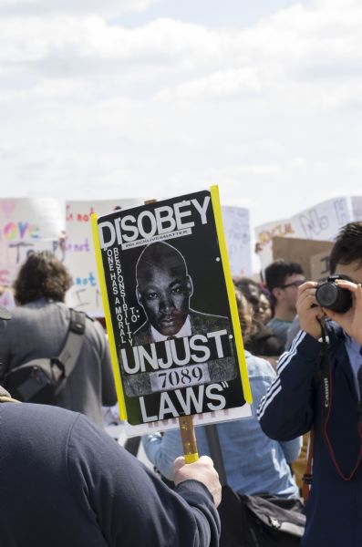 An individual holds up a sign which features the mug shot of Martin Luther King Jr. and a quote from him: "One has a moral responsibility to disobey unjust laws." Other protesters hold up their signs in the background and another person takes a photograph of the Martin Luther King Jr. sign.