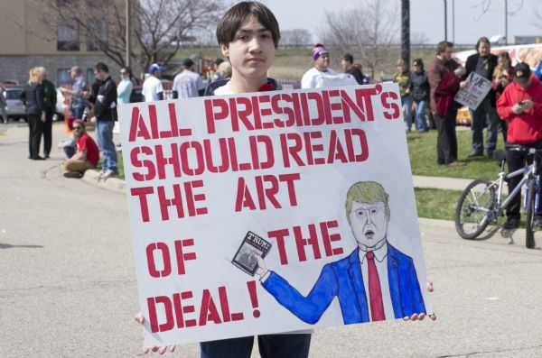 A young man is posing standing outdoors with his poster which reads: "All president's [sic] should read the Art of the Deal!" The sign features a drawing of Donald Trump holding up his book. There is a crowd of people standing on a lawn in the background.
