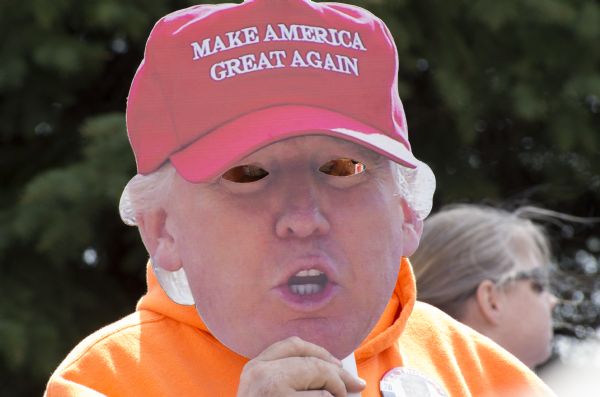 A Trump supporter is posing while holding up a mask of Donald Trump to cover their face. The mask features Trump wearing a red baseball cap with his campaign slogan: "Make America Great Again." The person is also wearing a Trump campaign button on their orange sweatshirt.