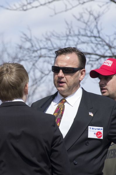 A man is standing with his son outdoors while waiting to enter the Donald Trump rally. They are both wearing suits, and the father is wearing sunglasses, a tie decorated with gray and gold leaves and red cherries, a pin of the American flag on his left lapel, and a square Trump political button on his breast pocket with a "I Voted" sticker attached to it. Another man standing behind him on the right is wearing a Packers shirt and a red cap with Trump's slogan: "Make America Great Again."