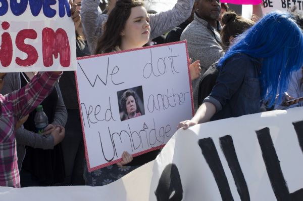 A young woman is holding up a protest sign standing among a crowd of men and women also holding signs. The woman's sign reads: "We don't need another Umbridge." It features a photograph of the character Umbridge from the <i>Harry Potter</i> movies. In the foreground a person is helping to hold up a section of a banner.