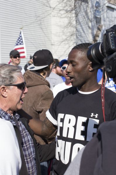 Ken Nwadike, founder of the Free Hugs Project, is wearing a black and white "Free Hugs" t-shirt while starting to embrace an elderly man. A man in the foreground is filming them with a camera. One man in the background is carrying a small American flag.