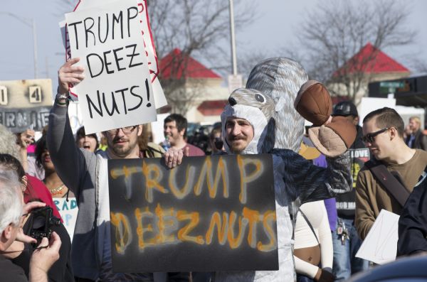 Two men hold up protest signs, both reading "Trump deez [sic] nuts!" The man on the right wears a full body squirrel costume and holds a pair of cloth acorns. They stand in front of a crowd of protesters. In the bottom left corner a man takes a picture of the two.