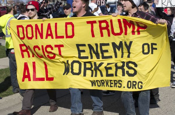 Three men walking and holding a banner which reads: "Donald Trump racist enemy of all workers.workers.org," in front of a crowd of protesters outside the Donald Trump rally.