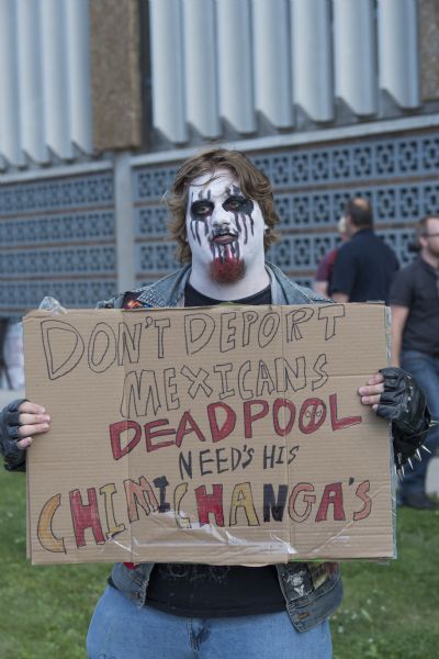 A man posing standing outdoors with his protest sign. He is wearing white, black, and red face paint. His sign reads: "Don't Deport Mexicans, Deadpool Need's [sic] his Chimichanga's [sic]."