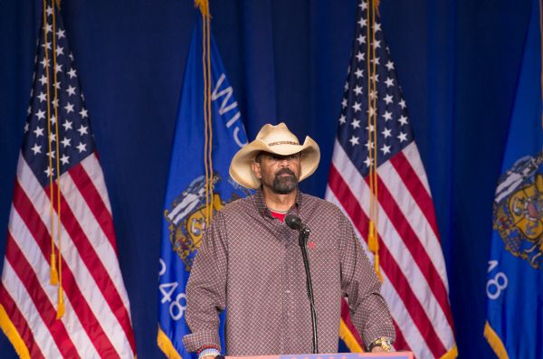 David A. Clarke Jr., Sheriff of Milwaukee County, standing at a podium ready to address the audience at the Donald Trump rally. He is wearing a plaid button shirt and a tan cowboy hat. Behind him are two American flags and two Wisconsin flags.