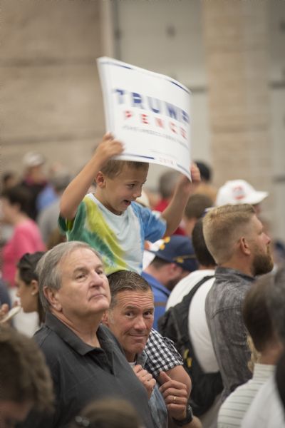 A father is holding his young son on his shoulders in a crowd of supporters at a Donald Trump rally. The boy is smiling and holding up a Trump/Pence political poster.