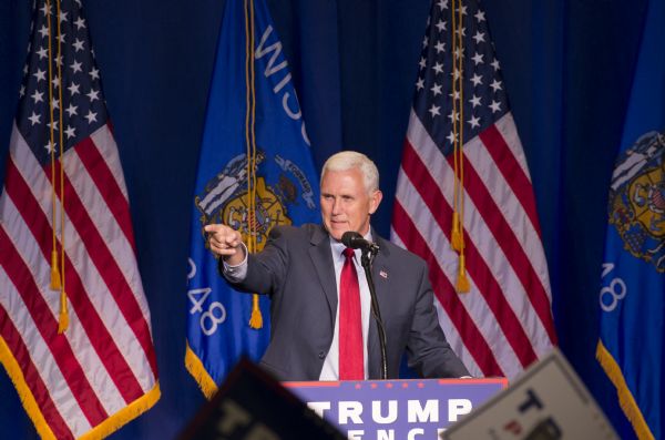 Governor of Indiana, and Donald Trump's running mate, Mike Pence, standing behind a podium. He is pointing at the audience and is wearing a suit, red tie, and an American flag pin on his lapel. Behind him are two American flags and two Wisconsin flags.