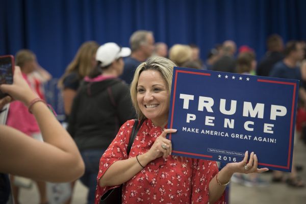 A woman is smiling and posing with her Trump/Pence campaign sign as another woman, obscured on the left, is taking her picture with a cell phone.