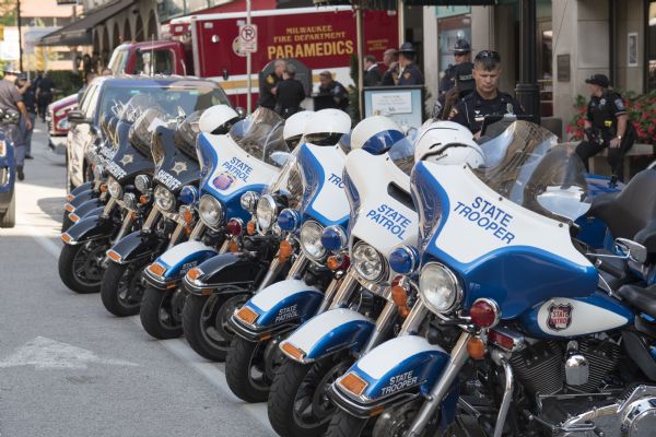 A line of motorcycles parked along a street, all marked as belonging to the state trooper or the sheriff department. At the end of the row of motorcycles is an ambulance backed up into an alley. Police officers and troopers are standing on the sidewalk.