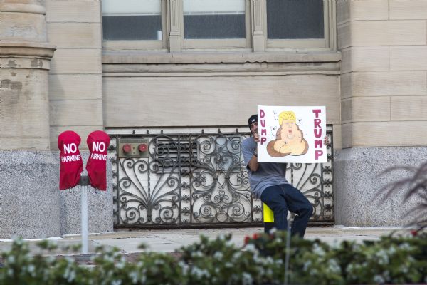 A man is sitting on a sidewalk in front of a building holding up his protest sign, near the Trump rally, which reads: "Dump Trump." and includes a hand-drawn caricature of Donald Trump surrounded by flies. 