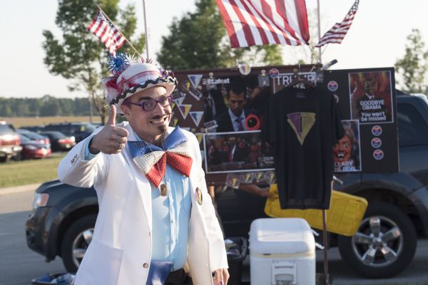 A man is posing outdoors in front of a display of his merchandise in a parking lot. He is smiling and giving a thumbs up, and is wearing a white suit jacket over a pale blue button-up shirt with an oversized red, white and blue bow tie. His bowler hat is mostly white but decorated with red, white, and blue feathers, ribbons, pom-poms, and a barbed wire motif along the rim of the hat. His eyeglasses are purple. His mustache and goatee are waxed into fine points. The merchandise in the background displays pins, stickers, and shirts, and almost all bear the same symbol: a red and gold upside down triangle with a 'T' in the middle. There are also pro-Trump, and anti-Hillary and anti-Obama posters.