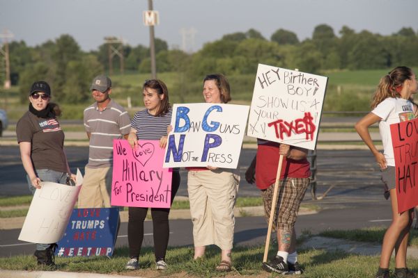 A line of men and women standing in a parking lot holding up their protest signs. One calls for Donald Trump to release his tax records, another is in support of Hillary Clinton. Most of the signs read: "Love Trumps Hate." A woman in the center is holding up a sign reading: "Ban Guns Not The Press."