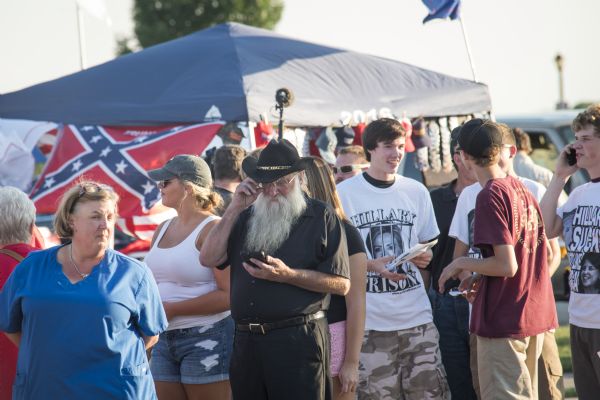 A crowd of people waiting for the start of the rally. A group of young men are wearing anti-Hillary t-shirts. Behind the group a tent is displaying merchandise for sale, including a Confederate flag.