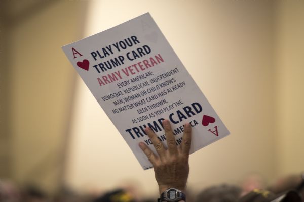 View of an arm holding up a poster with the design of a large playing card, with the ace of hearts in the upper left and bottom right corner. The poster reads: "Play Your Trump Card Army Veteran. Every American: Democrat, Republican, Independent, man, woman or child knows no matter what card has already been thrown, as soon as you play the Trump card you and America win!"
