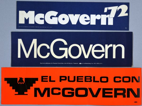 Three bumper stickers. The top bumper sticker has white text on a blue background and reads: "McGovern'72." The middle bumper sticker has white type on a blue background and reads: "McGovern." The bottom bumper sticker has a southwestern style eagle shape, and black letters on an orange background that reads: "El Pueblo Con McGovern."