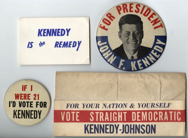 An assortment of four political campaign items for John F. Kennedy. At top left is a printed sign with blue text on white that reads: "Kennedy is the Remedy." Top right is a button in red, white and blue with a head and shoulder portrait of Kennedy that reads: "John F. Kennedy for President." At bottom left is a button that reads: "If I Were 21 I'd Vote for Kennedy." At bottom right is a white paper hat with red, white and blue text that reads: "For Your Nation & Yourself Vote Straight Democractic Kennedy-Johnson."