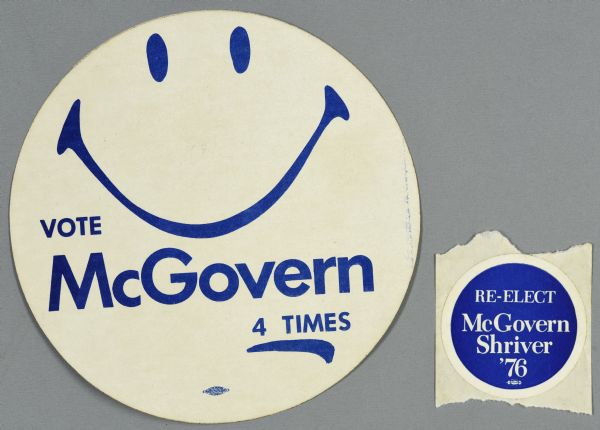 Two presidential political campaign stickers for George McGovern. The sticker on the left is white with blue text and displays a smiley face and reads: "Vote McGovern 4 times." The sticker on the right is blue with white text and reads: "Re-elect McGovern Shriver '76."