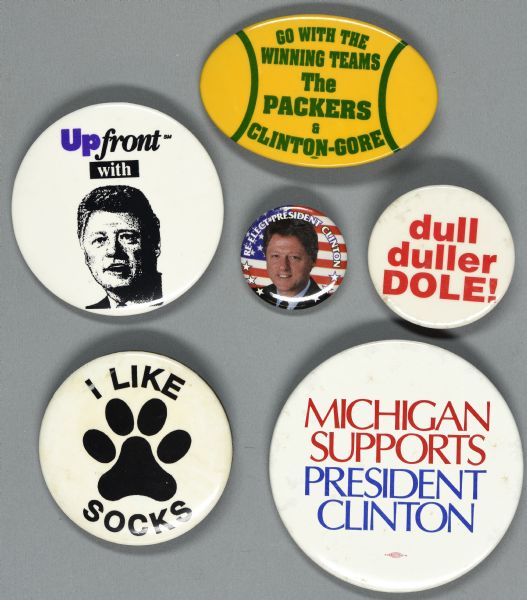 Six assorted presidential political campaign buttons for Bill Clinton, Al Gore and Bob Dole. One button is green and yellow (or gold), and has an oval shape, mimicking the shape of an American Football. It reads: "Go with the winning teams, The PACKERS & CLINTON-GORE." Another button is white with red text and reads: "dull, duller DOLE!" 
