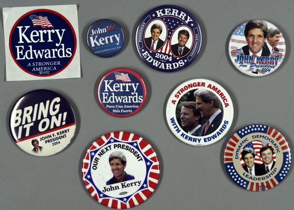 An assortment of presidential political campaign buttons and stickers for John Kerry and John Edwards. One  button uses gold and red stripes that alternate with blue and white stars to create a frame around a color head and shoulders portrait of John Kerry and John Edwards and reads: "Dynamic Democratic Leadership." Another button is circular and is split in half by a background from dark blue to white. It reads: "Bring it On! John F. Kerry President 2004."