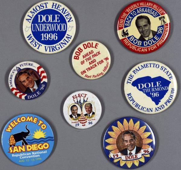 Eight assorted presidential political campaign buttons. Seven of them are for Bob Dole, and one is for the 1996 San Diego Republican National Convention, which is light blue with an image of an elephant wearing flower-printed swimming trunks, next to a sun and water. One button is dark blue with an image of a sunflower, with a portrait of Bob Dole in the center, which reads: "19 DOLE 96." Another button has a blue circle in the center with white text within and reads: "DOLE Underwood 1996," and blue text is on the border around the blue circle and reads: "Almost Heaven West Virginia!"
