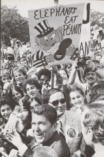 Slightly elevated view of Ronald Reagan standing outdoors among a crowd of young people and shaking hands. Someone in the crowd is holding a large sign that has a caricature of an elephant, wearing a stars and stripes hat and jacket, eating a peanut with the face of Jimmy Carter on it, that reads: "Elephants Eat Peanuts." On the back is pre-printed text that reads, in part: "Dear _______, I want to let you know that I am one of the many thousands of young people who are working hard to elect Ronald Reagan President of the United States." 