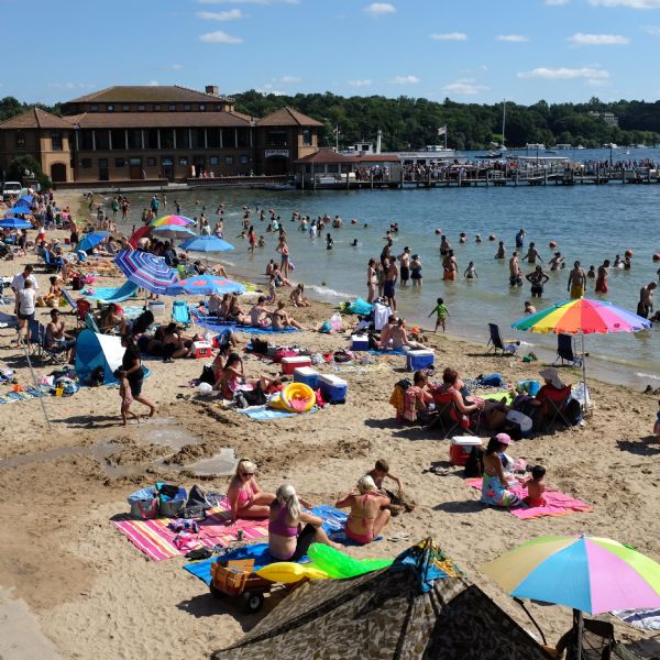 Elevated view of adults and children on the sandy beach of Geneva Lake. Many of the people are sitting on colorful beach towels, shaded by umbrellas. Other people are playing along the shoreline, or are in the water. The Riviera Ballroom is in the background, where a long line of people is waiting on the dock for a boat tour. Boats are at the docks and on the lake. Stone Manor is on the far shoreline among trees on the right.