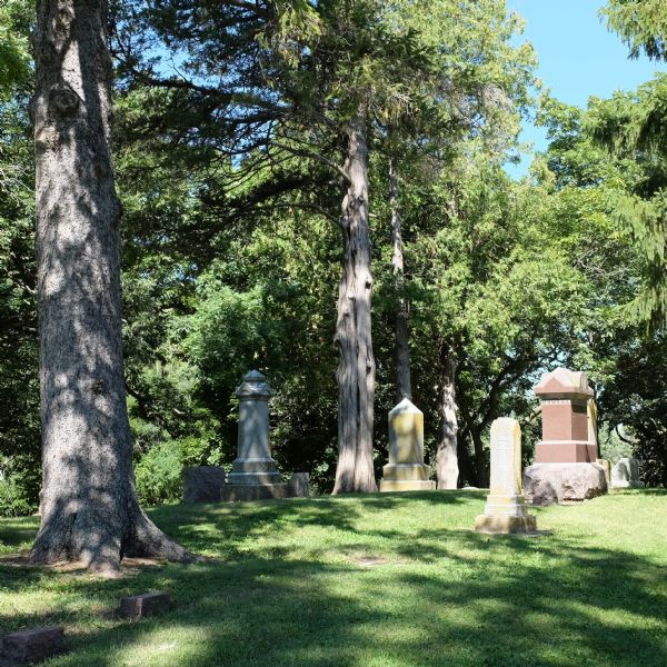 Large and small stone grave markers standing on a grassy lawn. Trees block the view of the lake below the cemetery.