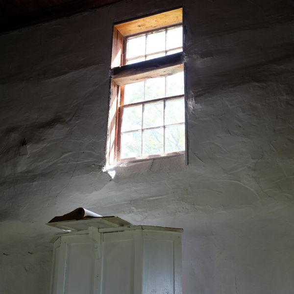 Interior view of a window sitting high up in a white lime-plastered wall, letting light into Hauge Church, a small Lutheran church built in 1852 by Norwegian immigrants. Below the window is a wooden pulpit on which an open book sits.