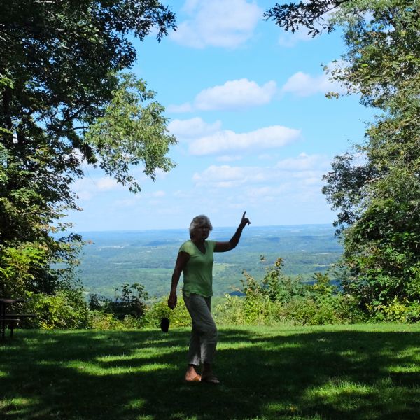 View from the top of a hill looking out to a valley, with clouds in the blue sky. Trees are framing the view, and in the foreground, a woman, the photographer's wife Solveig Quinney, is standing and pointing to the sky with her left hand.