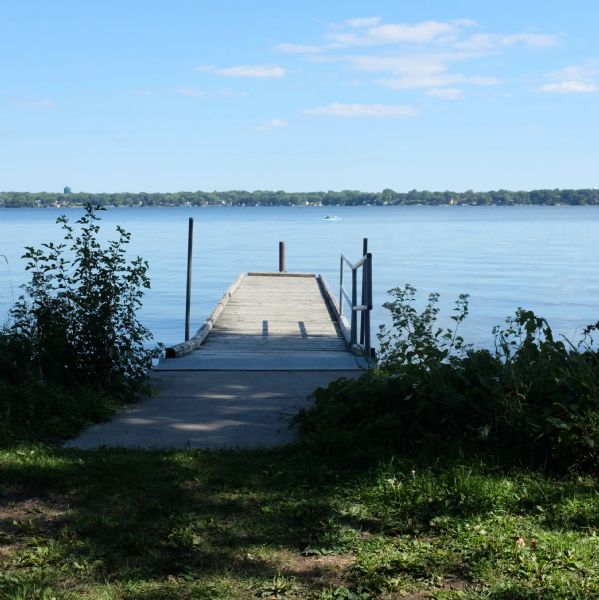 View from shoreline towards a small wooden pier on Lake Monona. Homes and buildings and a water tower are among trees on the opposite shoreline. There is a person paddling a blue kayak out on the lake.