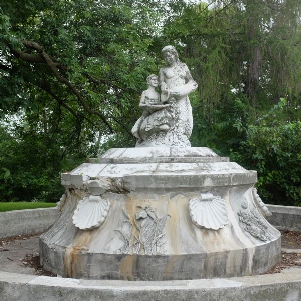 View of the Annie Stewart Memorial Fountain. The base of the circular fountain is decorated with a repeated motif of shells and flowers. A sculpture is at the top, depicting a mermaid pouring water into a conch shell held by the Greek god Triton, with a sea creature underneath them. The fountain is worn and water no longer runs from it. Trees are in the background. 