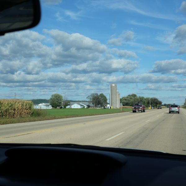 View through the windshield of a car towards several light blue farm buildings on the left side of a highway, along with three silo's. The edge of a field of corn is on the left. Cars and trucks are driving on the highway.