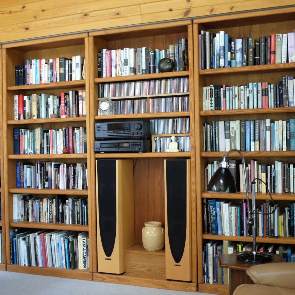 View of wooden bookshelves built into the wall. Books, cds, a cd player and radio, two tall speakers, small statues and pottery fill the shelves. A lamp on an end table is next to a leather chair in the right foreground.