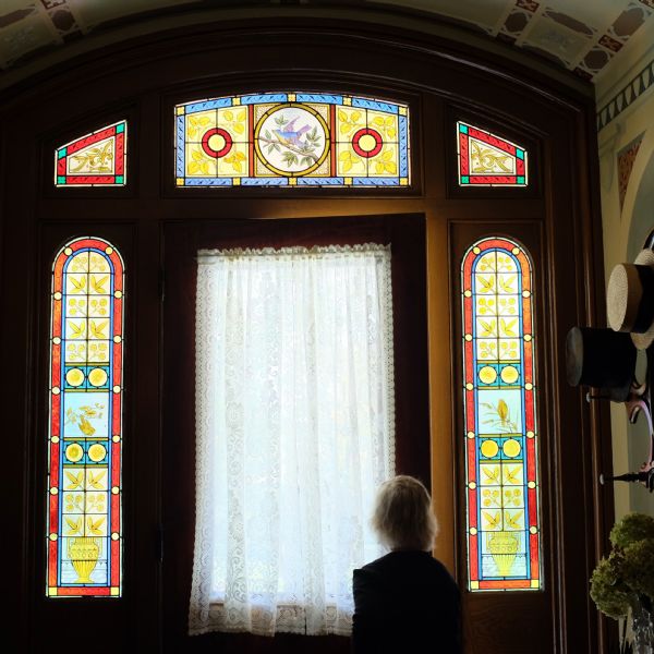 View of a woman (facing away) standing indoors in front of an ornate doorway at the Joseph Gundry House. A white lace curtain is covering the large window of the wooden door which is slightly ajar. There are five stained glass windows framing the door, including two sidelights with arched tops, and a three-part transom matching the arched shape of the ceiling. The design features birds, foliage, and Greek kraters. The walls and ceiling mimic the color motif of reds, blues and yellows in the stained glass. A hat rack with hats is against the wall on the right.