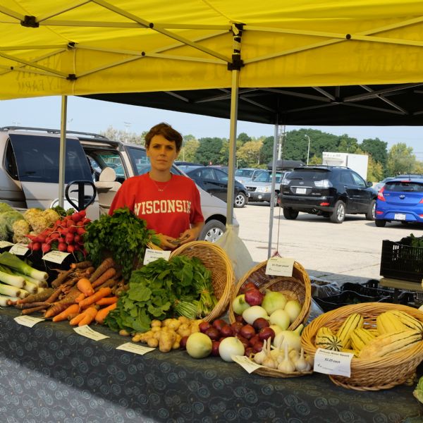 A woman is standing behind a table with a display of vegetables. She is wearing a red shirt with the word "Wisconsin" printed on it. The vegetables are arranged in baskets or on stands with labels indicating their names and prices. A yellow tent with the words "JenEhr Family Farms" on the side is shading the stand. Cars are parked in the parking lot in the background.