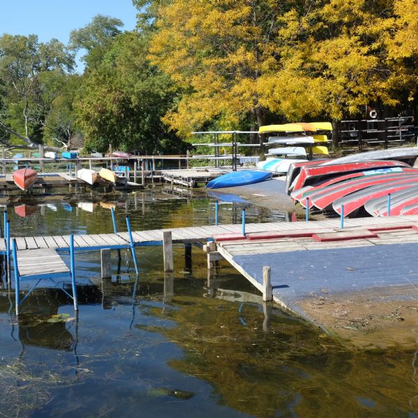 View across water towards canoes and kayaks stored on the sand, on docks, or on berths on the shoreline of Lake Wingra.