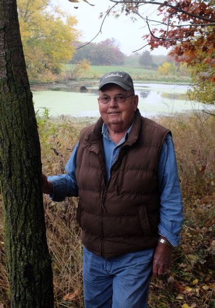 Portrait of Ralph Quinney, standing with his right hand on the trunk of a tree. He is wearing eyeglasses, and a baseball cap with the word "Cabela's" written on it, jeans, and a brown vest. In the background is the pond surrounded by trees and plants turning fall colors.